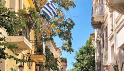 A picture of a street in Plaka, Athens taken on a sunny day. There are residential homes with balconies, plants and a Greek flag.