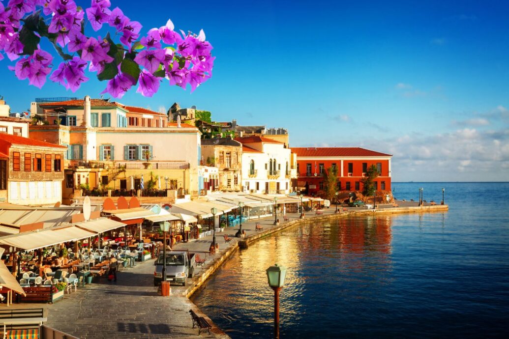 A picturesque view of Chania old town in Crete, featuring colorful buildings along the waterfront, outdoor cafes, and a calm, blue sea under a clear sky with vibrant purple flowers in the foreground.
