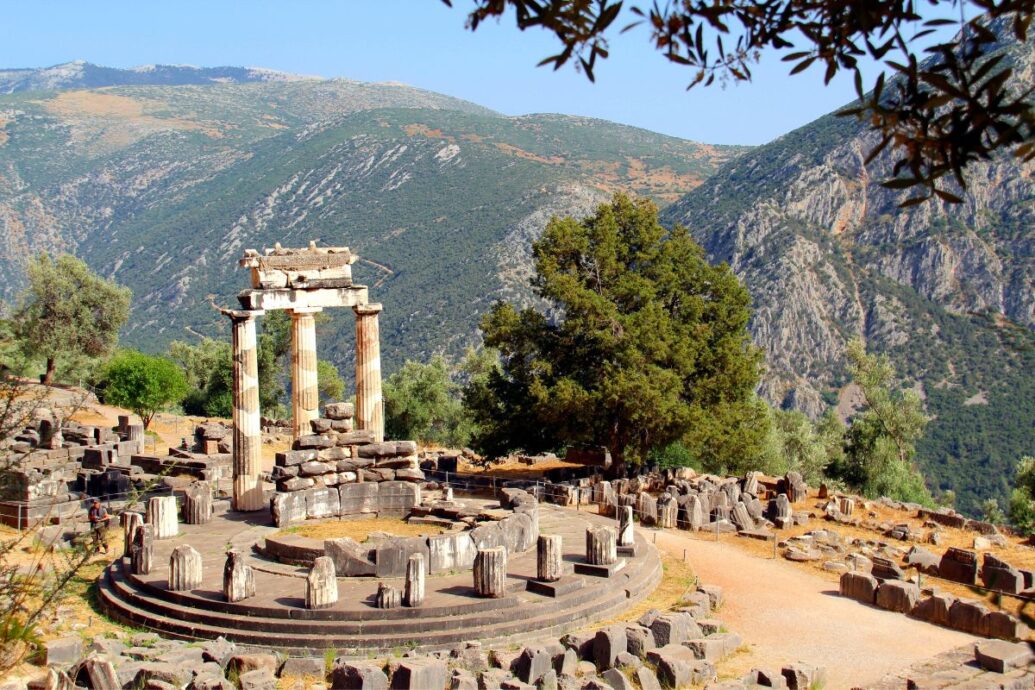 The image shows the ruins of the Tholos of Delphi, with partially standing columns, surrounded by trees and mountains in the background.