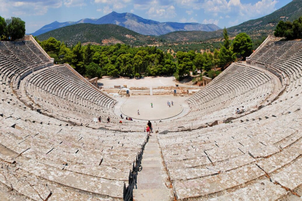 The image shows the ancient theater of Epidaurus, featuring large stone seating arranged in a semi-circle, with a central stage area and a backdrop of trees and mountains.