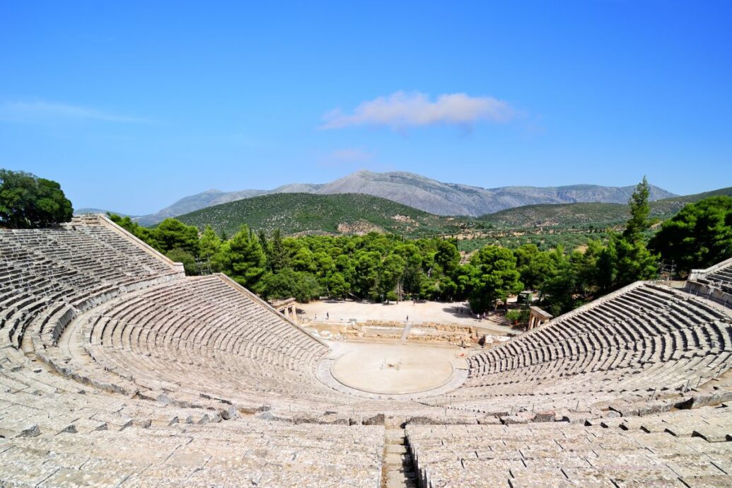 The ancient theater of Epidaurus, surrounded by lush greenery and mountainous landscape, features stone seating arranged in a semi-circular layout, showcasing its architectural brilliance and historical significance.