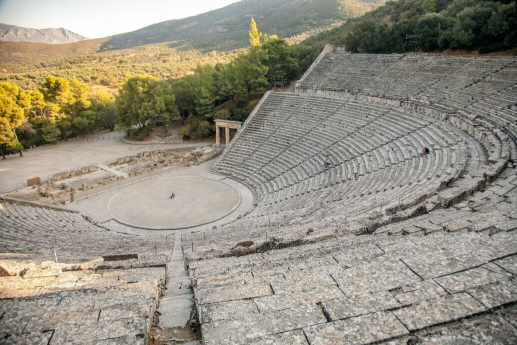 A large ancient amphitheater, Epidaurus in Greece, partially surrounded by lush green hills, showcasing the grand stone seating and central performance area.