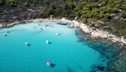 An aerial view of Halkidiki, showcasing clear turquoise waters, a rocky shoreline, and several small boats floating near the coast, surrounded by lush green trees.