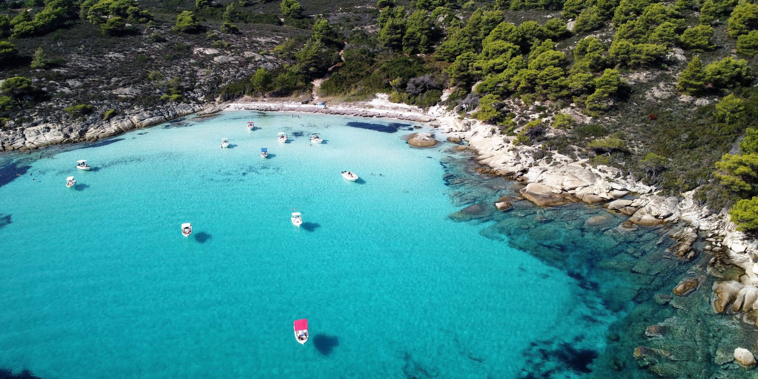 An aerial view of Halkidiki, showcasing clear turquoise waters, a rocky shoreline, and several small boats floating near the coast, surrounded by lush green trees.