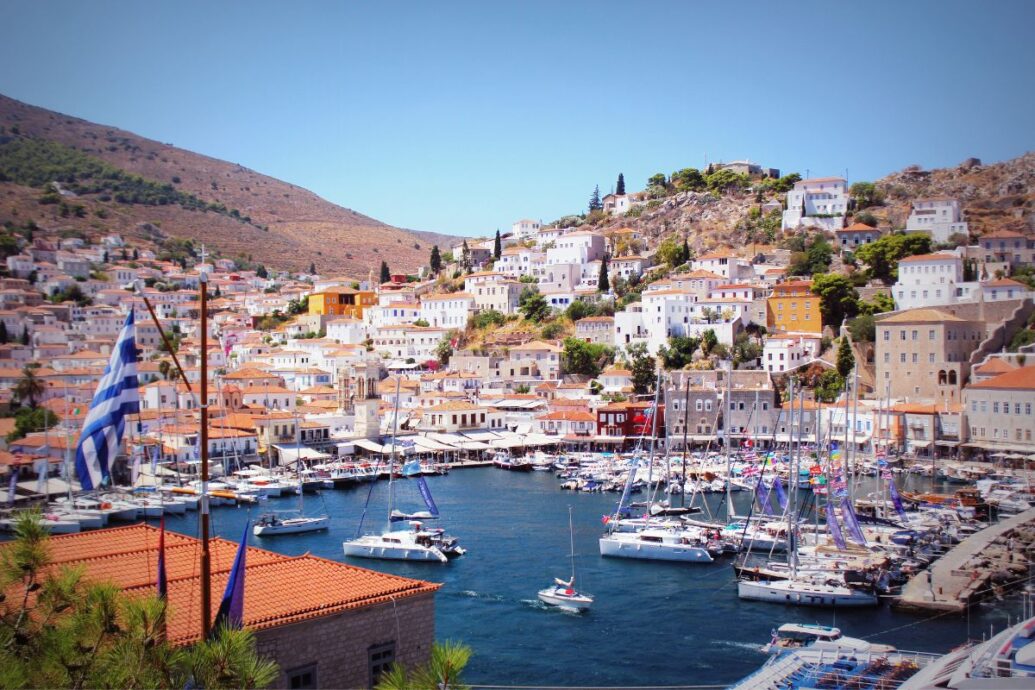 The image shows Hydra Island, featuring a picturesque harbor filled with boats, surrounded by white and pastel-colored houses built on a hillside. A Greek flag is visible in the foreground.