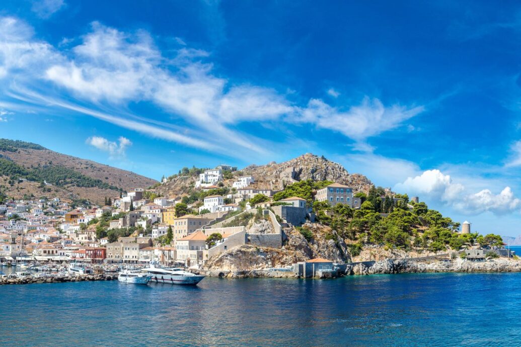 A picturesque view of Hydra showcasing traditional houses on the hillside and boats docked at the harbor, set against a backdrop of clear blue skies and azure waters.