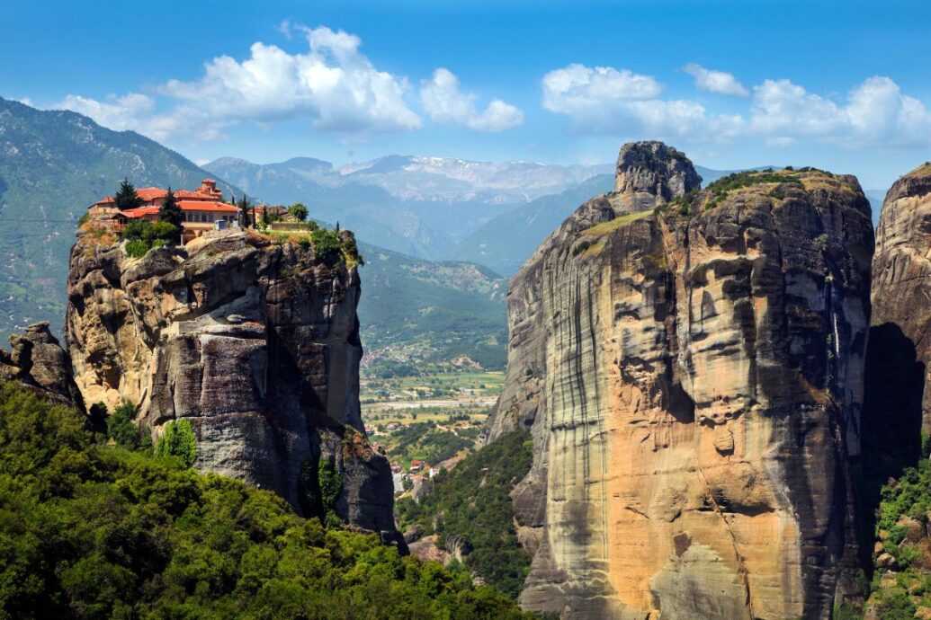 A panoramic view of Meteora, featuring a monastery perched atop a towering rock formation, surrounded by lush green landscapes and distant mountains under a bright blue sky.