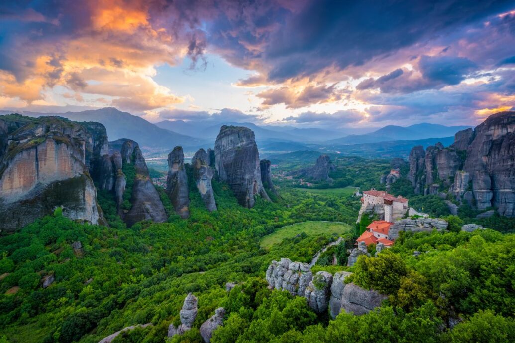 A stunning view of Meteora in Greece, showcasing dramatic rock formations and monasteries perched atop the cliffs, with lush greenery and a vibrant sunset sky in the background.