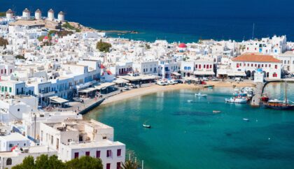 A panoramic view of Mykonos, showcasing the charming whitewashed buildings, windmills on the hillside, and a vibrant harbor with boats, all set against the backdrop of the deep blue Aegean Sea.