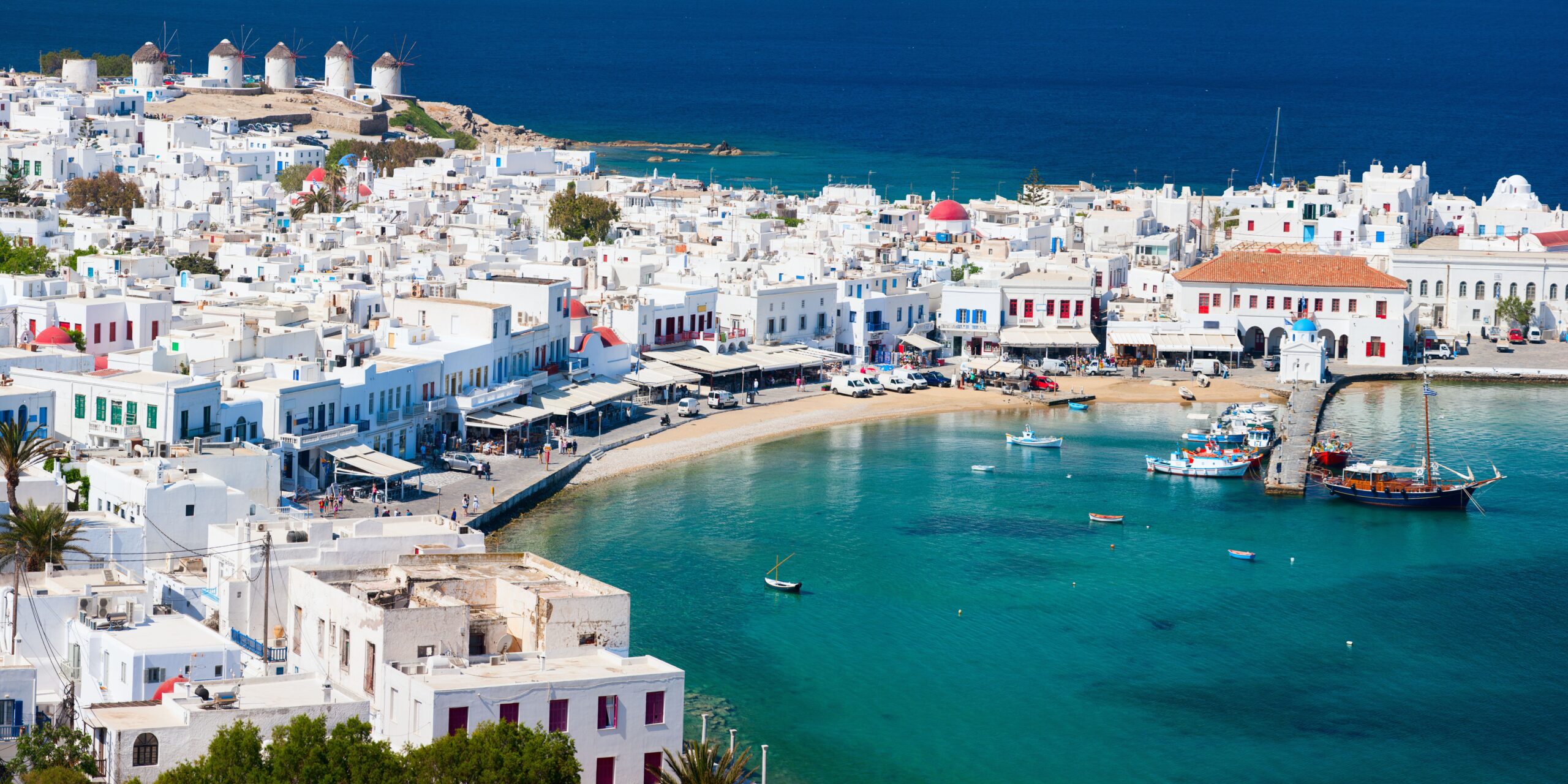 A panoramic view of Mykonos, showcasing the charming whitewashed buildings, windmills on the hillside, and a vibrant harbor with boats, all set against the backdrop of the deep blue Aegean Sea.