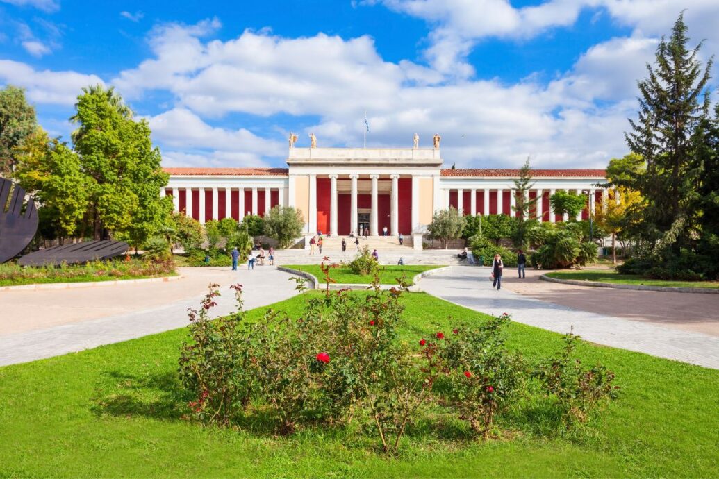 The image shows the National Archaeological Museum in Athens, featuring a grand building with red columns, statues on the roof, and a well-maintained garden in the foreground with pathways and visitors.