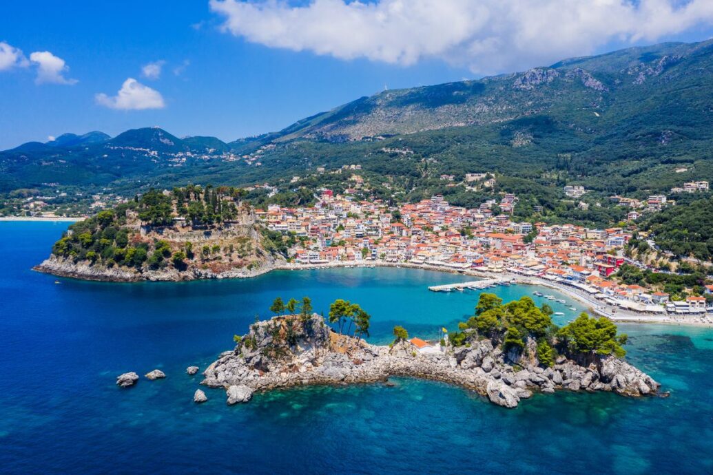 A scenic view of Parga town with its charming coastal architecture, vibrant buildings, and lush green hills surrounding the clear blue waters of the bay. The landscape is dotted with small boats and rocky outcrops.