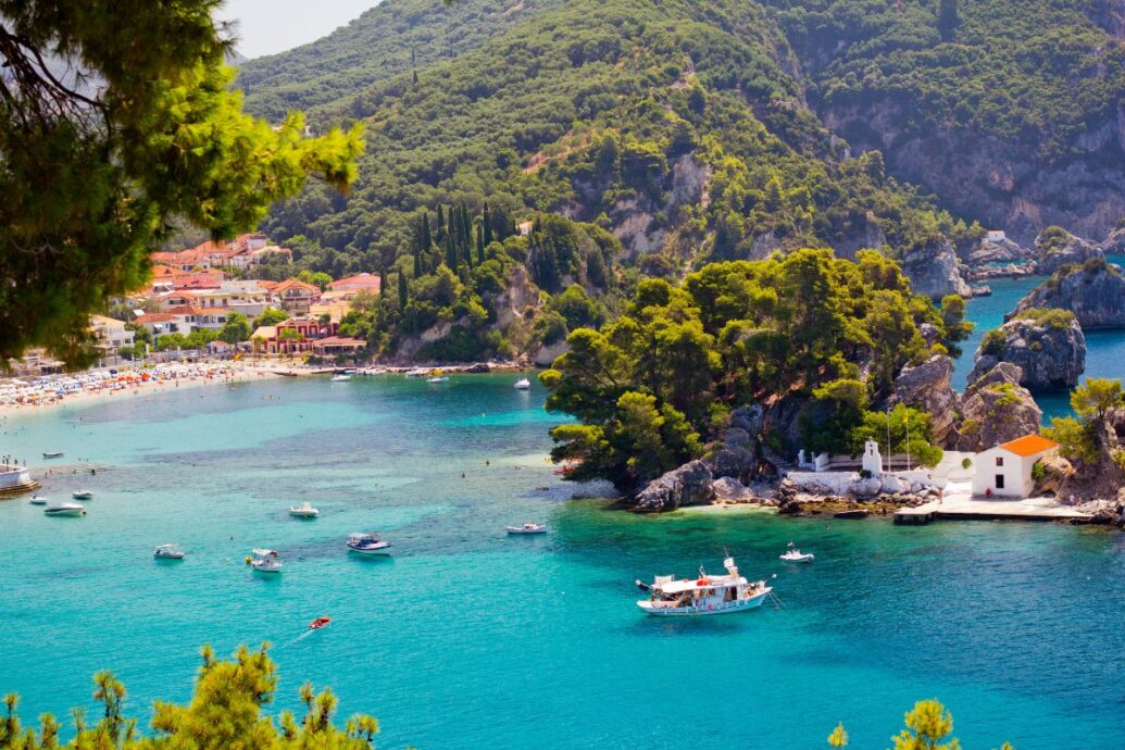 A picturesque view of Parga in Epirus, Greece, featuring turquoise waters, lush green hills, boats on the water, and a charming coastal village with colorful buildings and a small chapel by the shore.