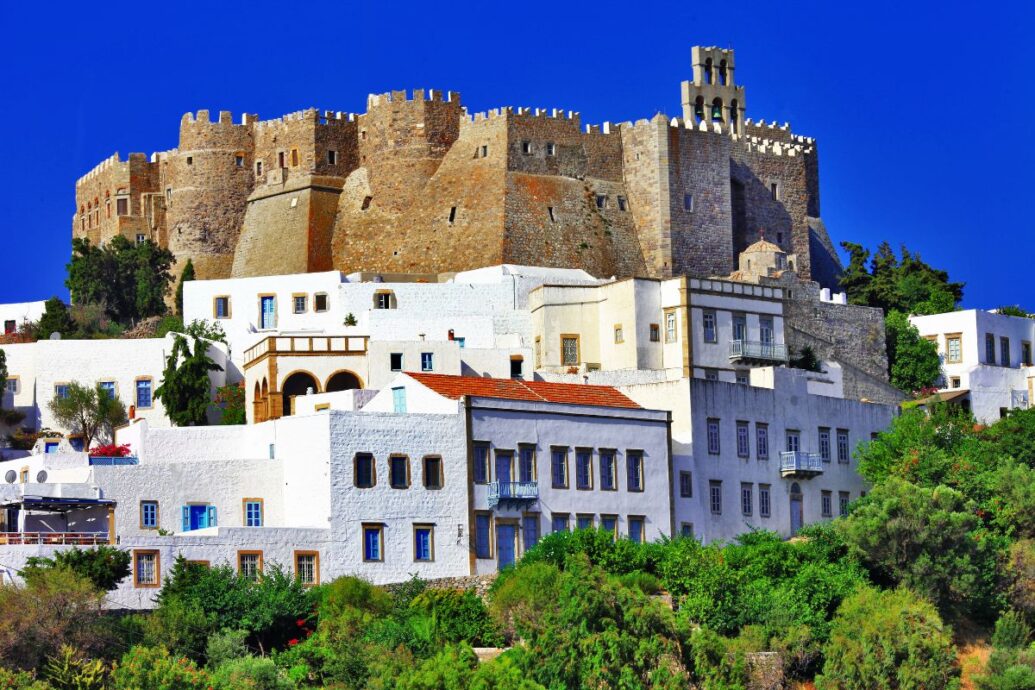 A view of Patmos Island featuring the imposing medieval castle with whitewashed buildings and lush greenery in the foreground under a clear blue sky.