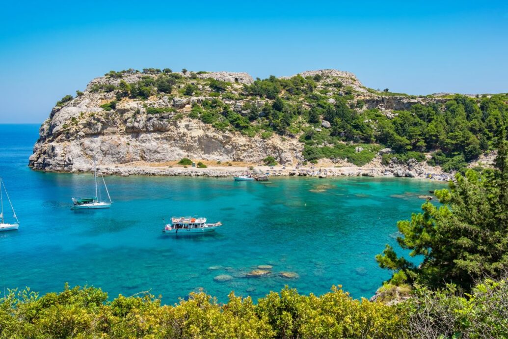 A picturesque view of Rhodes Island featuring clear turquoise waters, a rocky coastline, and lush greenery. Sailboats float peacefully in the calm sea, inviting a sense of serenity and adventure.