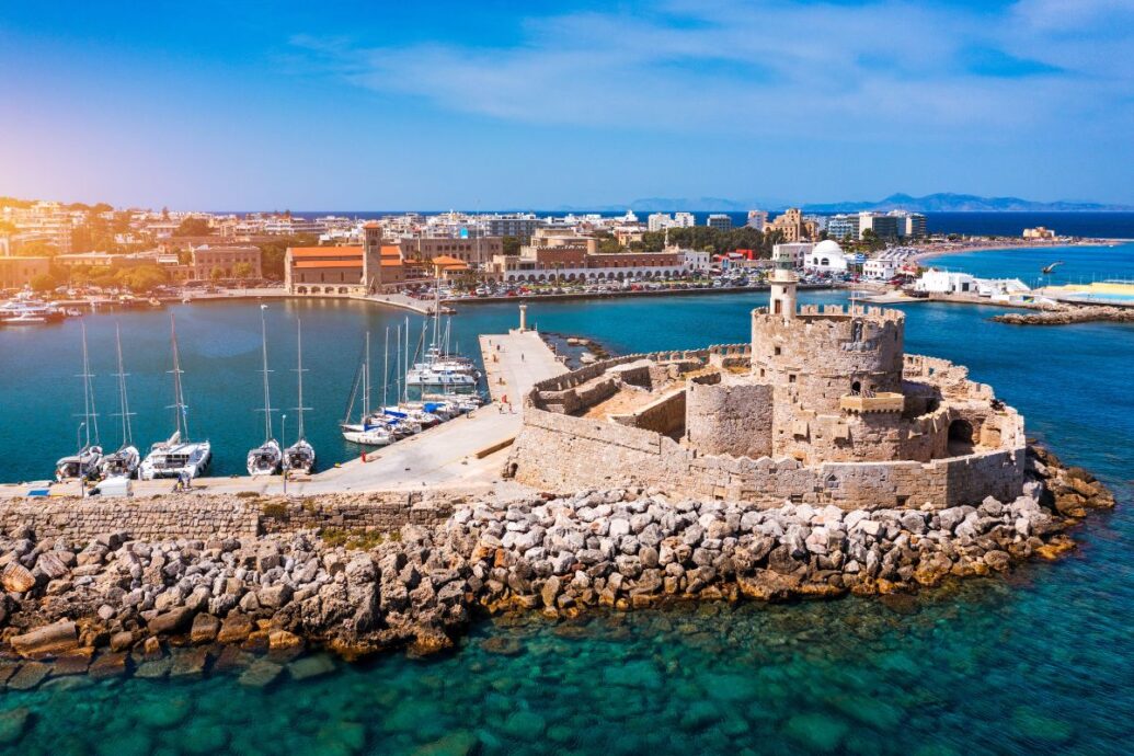 The image shows the harbor of Rhodes, featuring a medieval fortification on a rocky pier, surrounded by clear blue waters, with boats docked nearby and the town in the background.