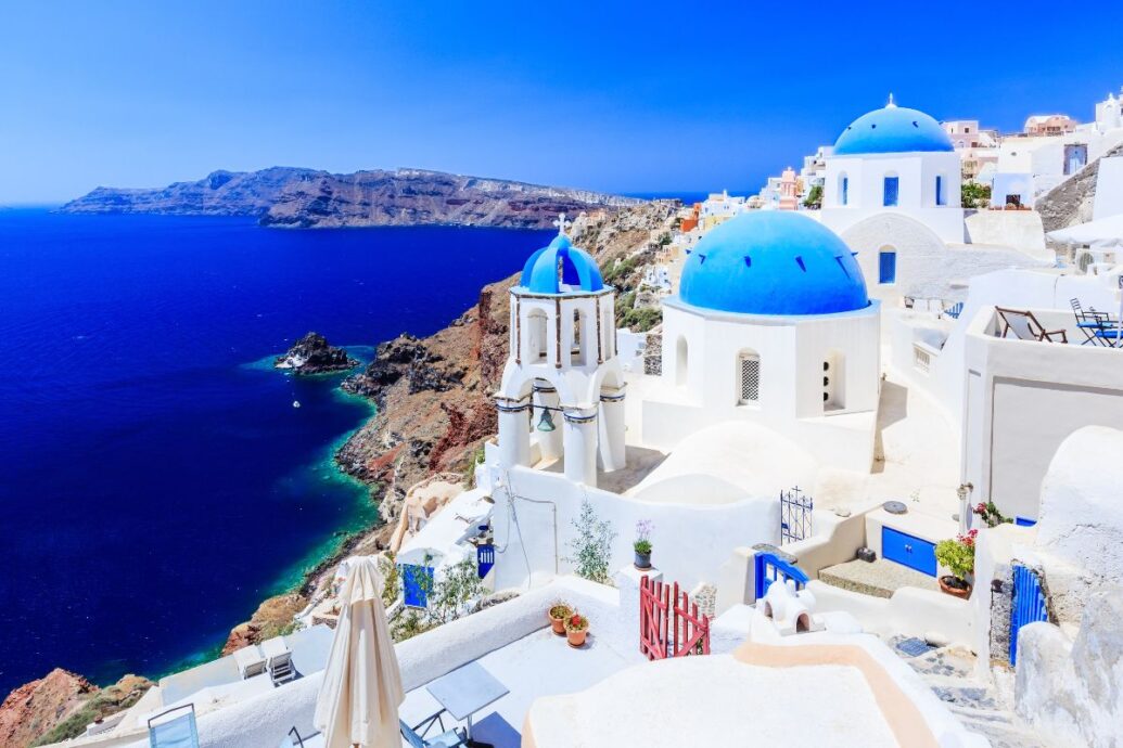 View of Santorini featuring iconic white buildings with blue domes perched on cliffs overlooking the deep blue Aegean Sea, with a stunning coastline in the background.
