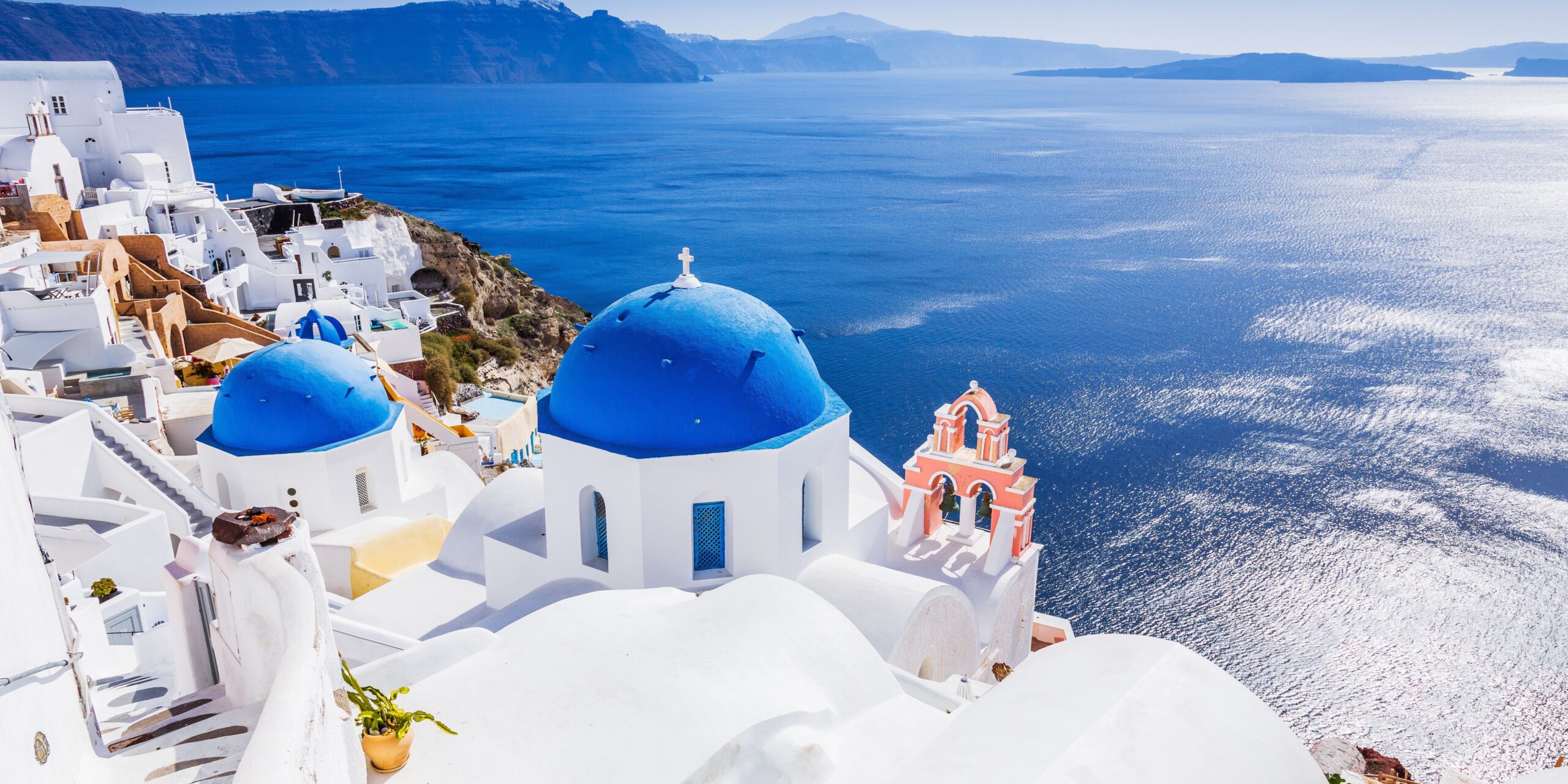 The image shows Santorini, featuring iconic white buildings with blue domes overlooking the deep blue Aegean Sea, with a rugged coastline in the background.