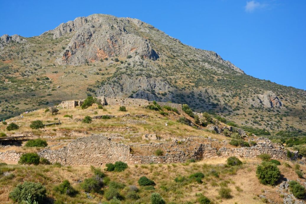 The image shows the ancient site of Mycenae, featuring stone ruins spread across a hillside with a rocky mountain backdrop under a clear blue sky.