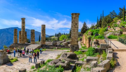 Tourists exploring ancient ruins at Delphi, Greece, with stone columns and a mountainous landscape in the background under a clear blue sky.