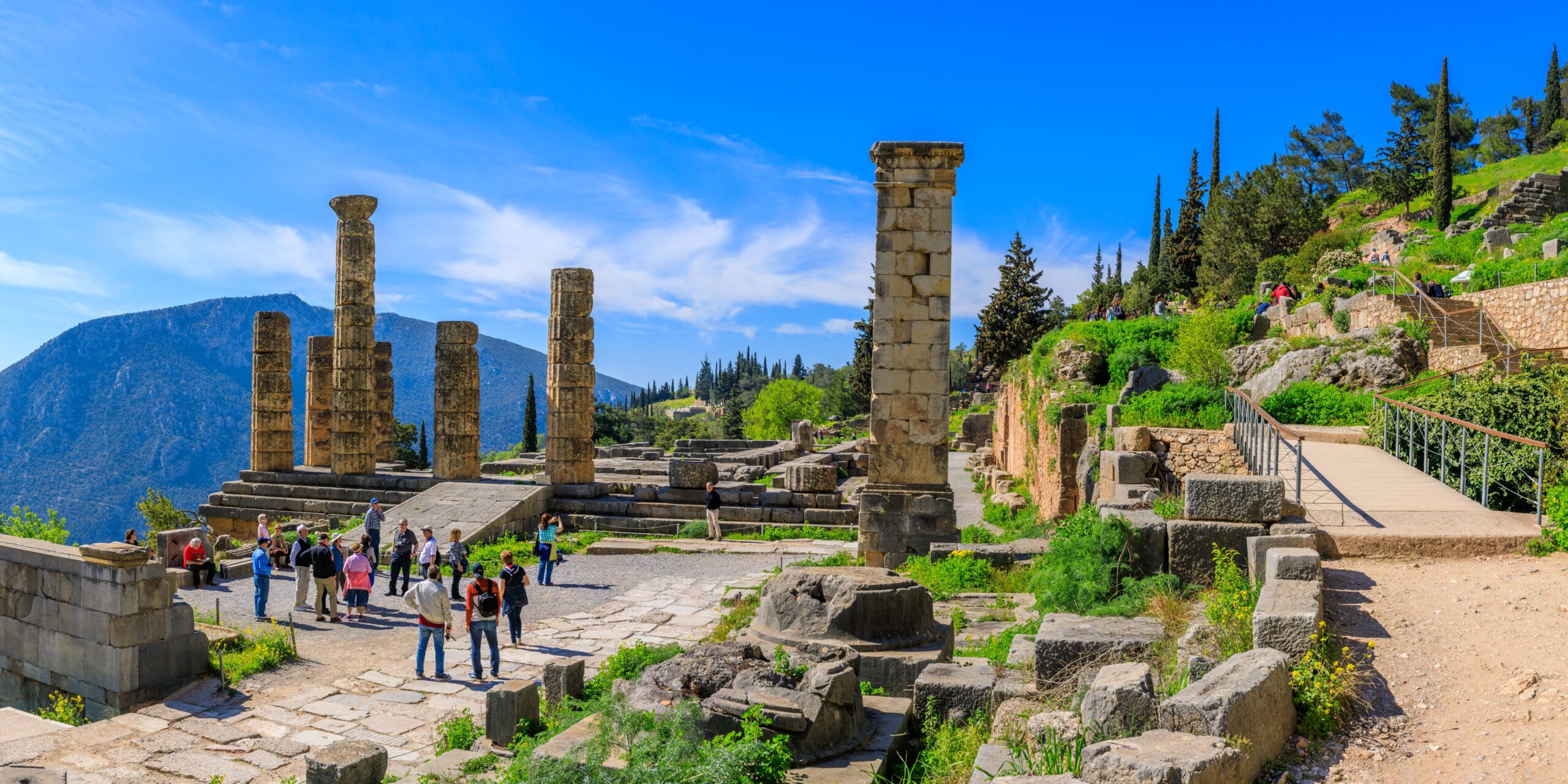 Tourists exploring ancient ruins at Delphi, Greece, with stone columns and a mountainous landscape in the background under a clear blue sky.