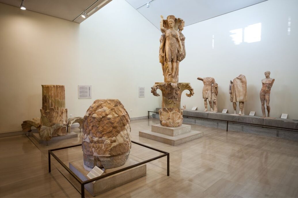 Interior of the Delphi Archaeological Museum, showcasing various ancient statues and artifacts, including a large column capital and a stone sculpture of the omphalos.