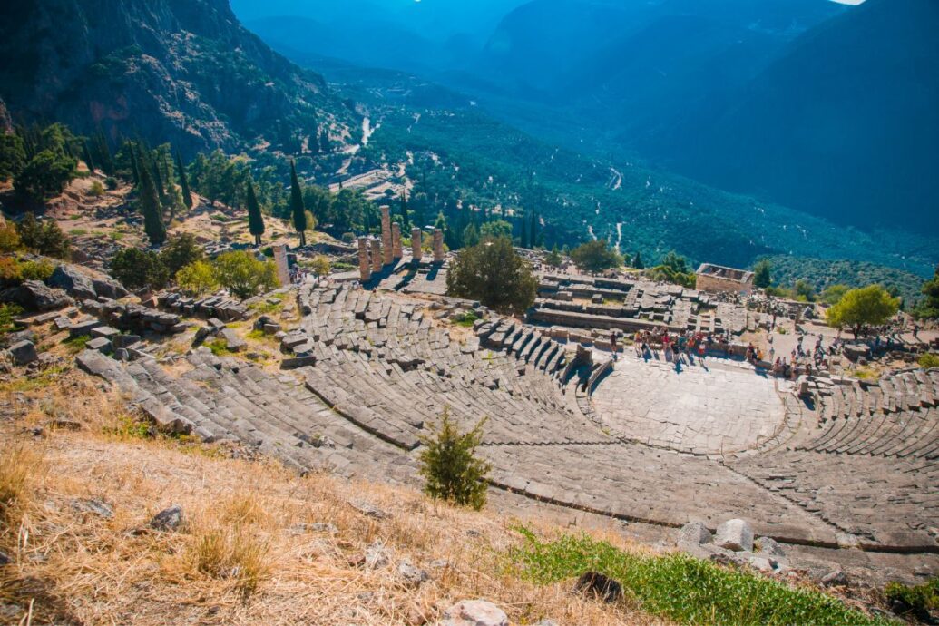 Ancient theater at Delphi, Greece, with stone seating and columns, overlooking a scenic valley and mountains in the background.