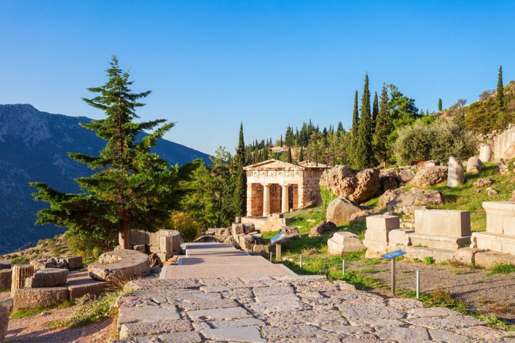 Pathway leading to ancient ruins at Delphi, Greece, with a small temple structure in the background, surrounded by greenery and mountains under a clear blue sky.