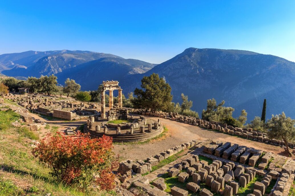 Ancient ruins of Delphi, Greece, featuring the Tholos of Delphi with surrounding mountains in the background under a clear blue sky.