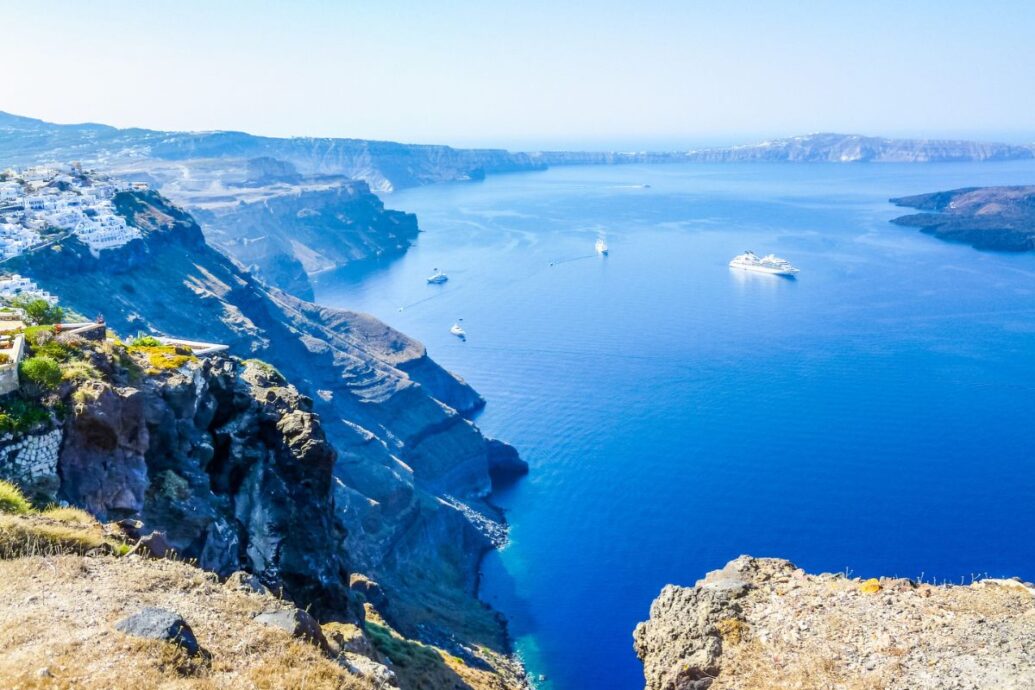 A stunning view of Fira in Santorini, featuring white buildings perched on steep cliffs overlooking the deep blue Aegean Sea, with boats and yachts dotting the water below.