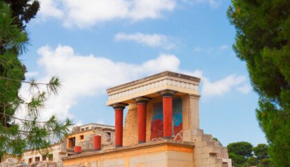 The image shows a section of the Knossos Palace, featuring reconstructed columns and vibrant frescoes under a bright blue sky with scattered clouds, framed by green trees on either side.