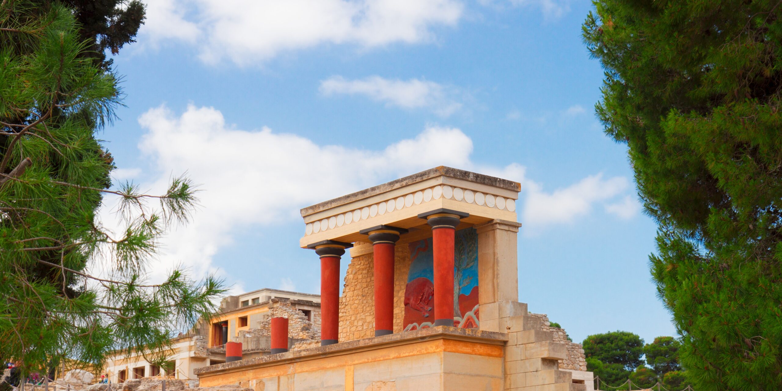 The image shows a section of the Knossos Palace, featuring reconstructed columns and vibrant frescoes under a bright blue sky with scattered clouds, framed by green trees on either side.