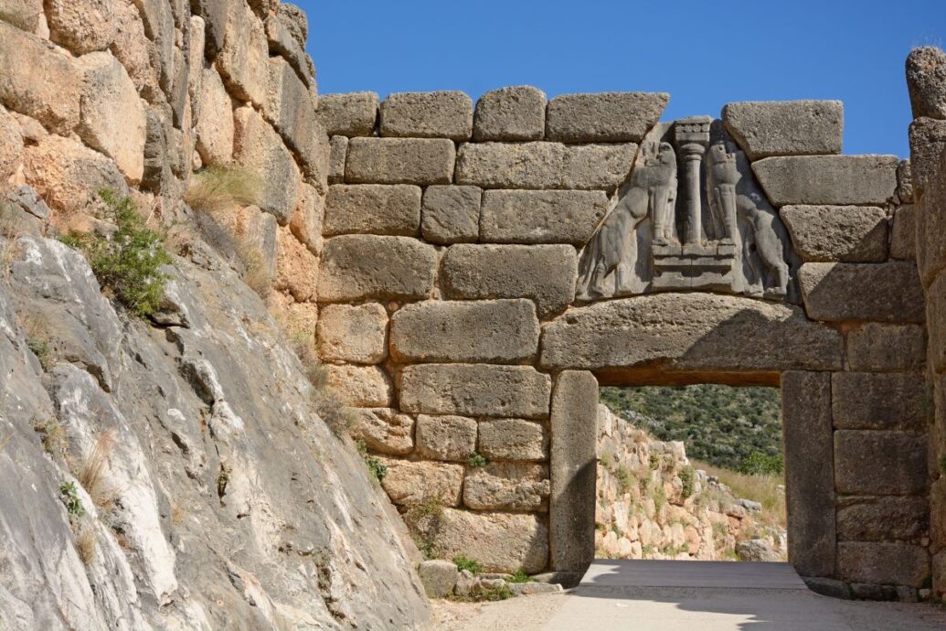 The image shows the Lion Gate at Mycenae, featuring large stone blocks and a carved relief of two lions above the entrance, under a clear blue sky.