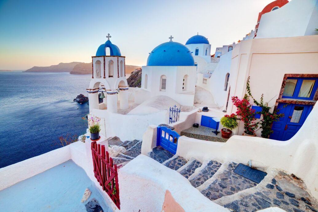 A charming view of Oia in Santorini, showcasing white-washed buildings with blue domes and picturesque stairways, overlooking the serene Aegean Sea at sunset.