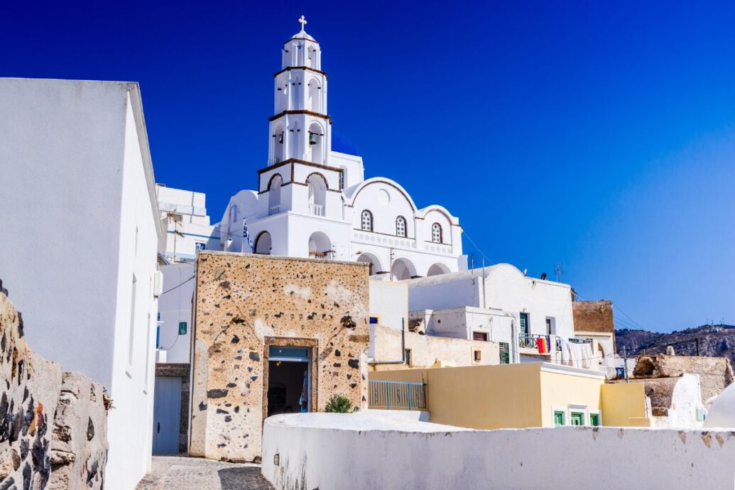 A view of Pyrgos Village in Santorini, featuring traditional white-washed buildings and a prominent church with a tall bell tower against a clear blue sky.