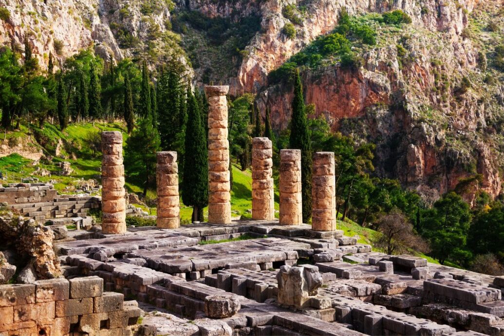 Ancient ruins at Delphi, Greece, featuring several standing stone columns with a backdrop of rocky cliffs and greenery.