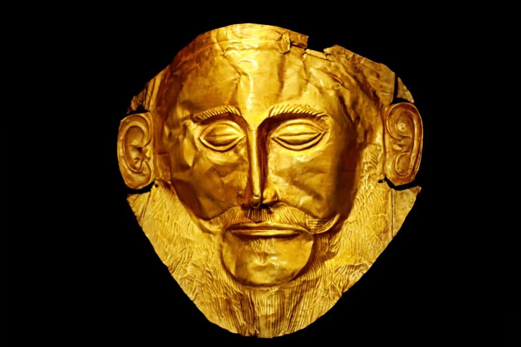 The image shows the golden Mask of Agamemnon, a funerary mask from Mycenae, depicting a bearded man's face.