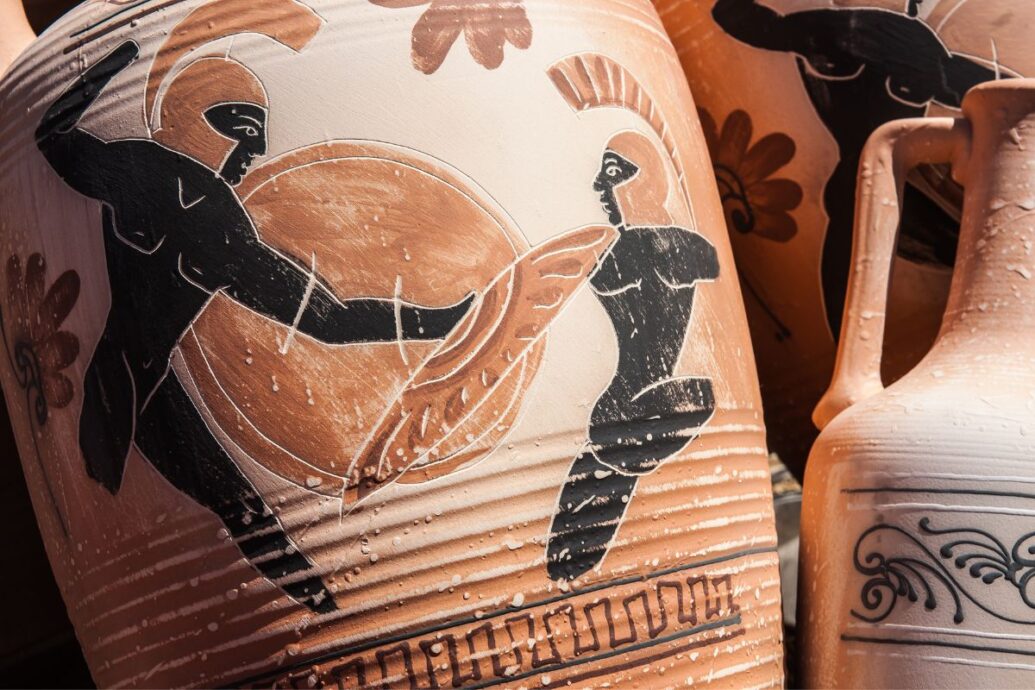 The image shows ancient Greek pottery decorated with black-figure paintings of warriors in combat, featuring detailed and intricate designs.