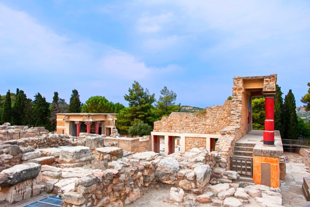 The image depicts the ruins of Knossos Palace, showcasing ancient stone structures and colorful reconstructed columns amidst a backdrop of trees and a blue sky.