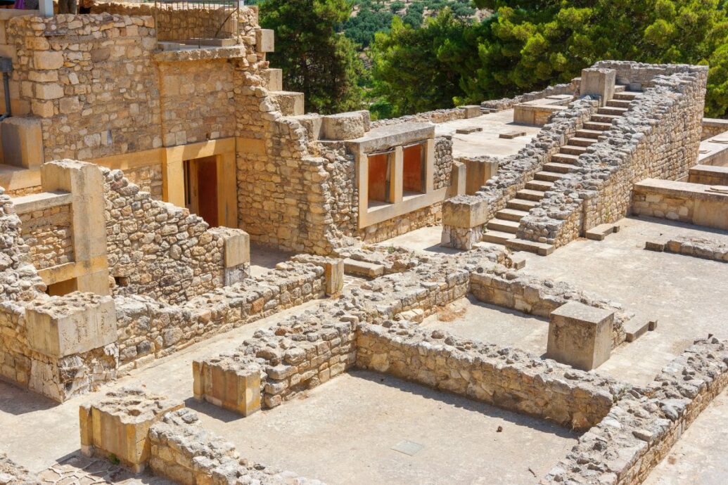 The image shows the archaeological site of Knossos Palace, featuring stone walls, staircases, and remnants of ancient structures amidst a lush green landscape.