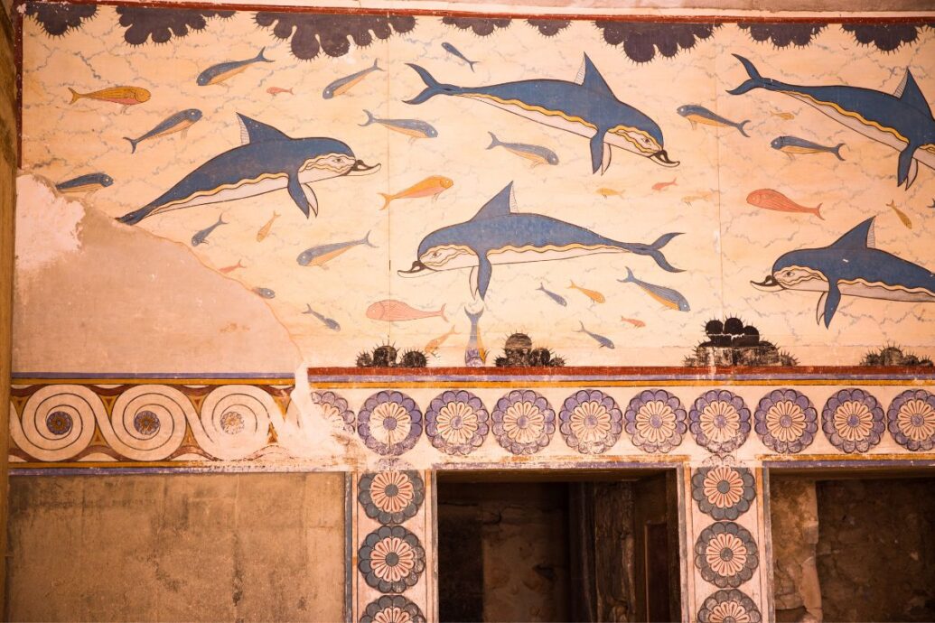 The image shows vibrant frescoes at Knossos Palace depicting dolphins and fish against a blue background, with intricate decorative patterns below.
