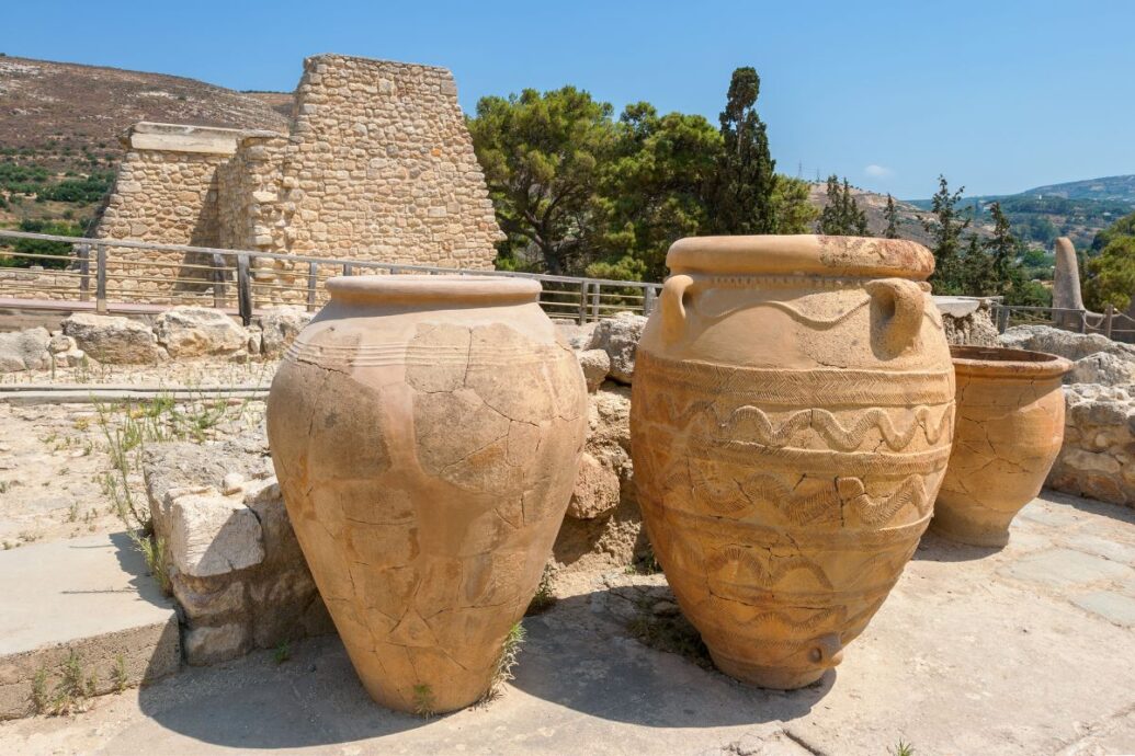 The image shows large Minoan pottery jars at Knossos Palace, with ancient stone walls and greenery in the background.