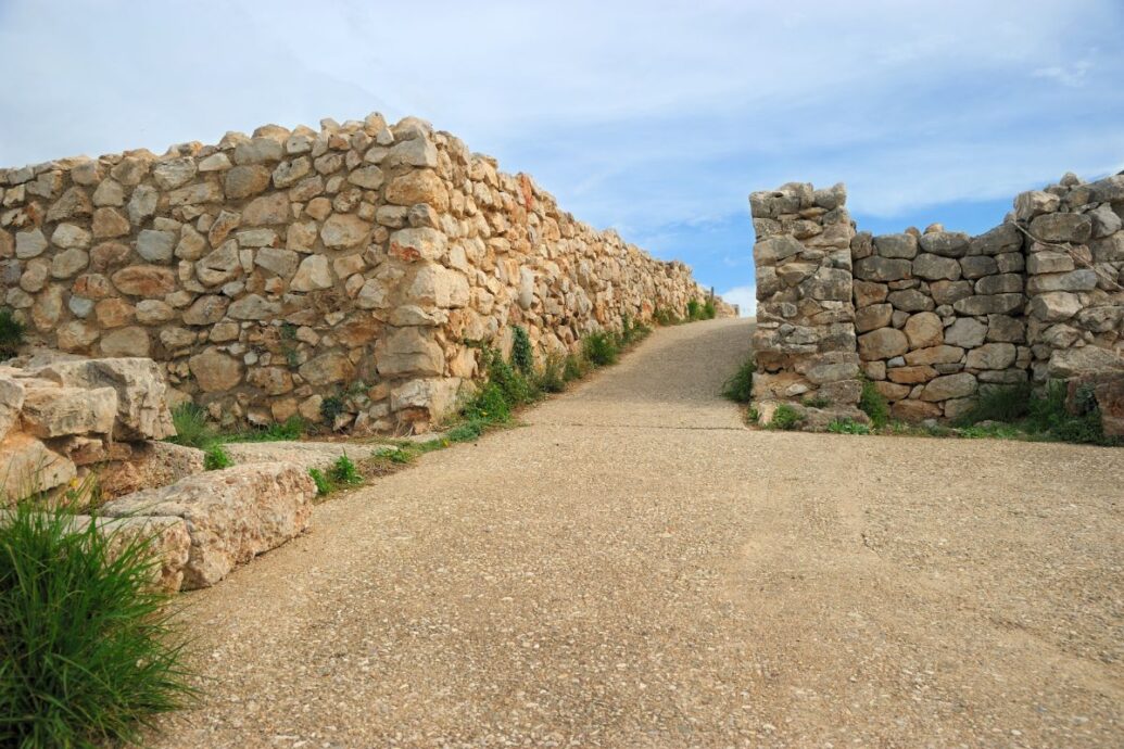 The image shows a pathway flanked by stone walls at the ancient site of Mycenae, under a clear sky.