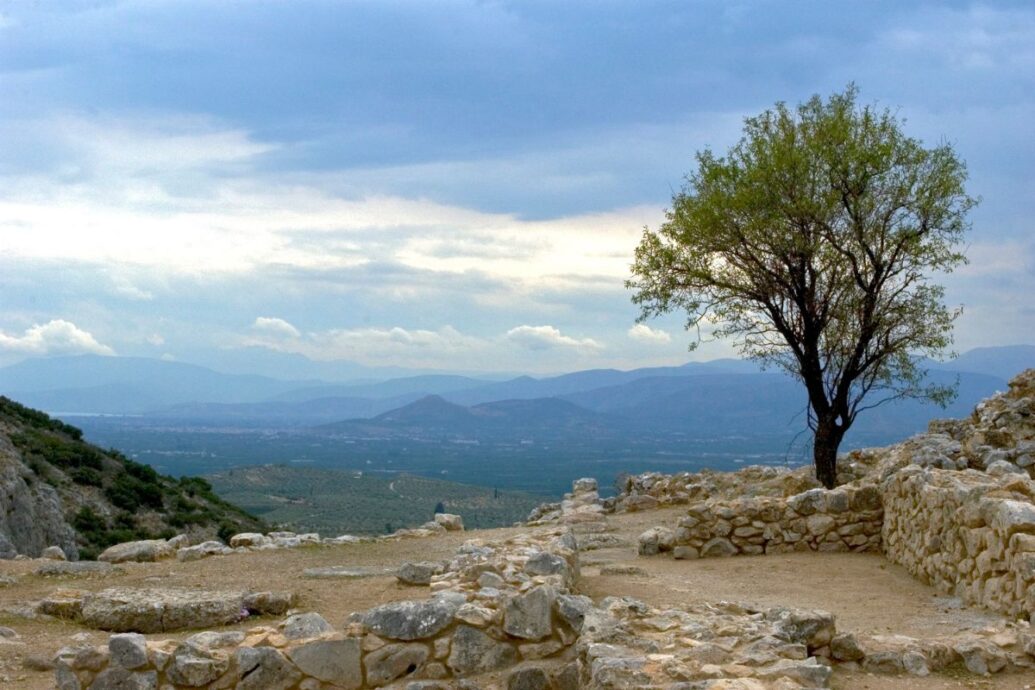 The image shows a view from the ancient site of Mycenae, featuring stone ruins in the foreground and a solitary tree, with a backdrop of rolling hills and a cloudy sky.