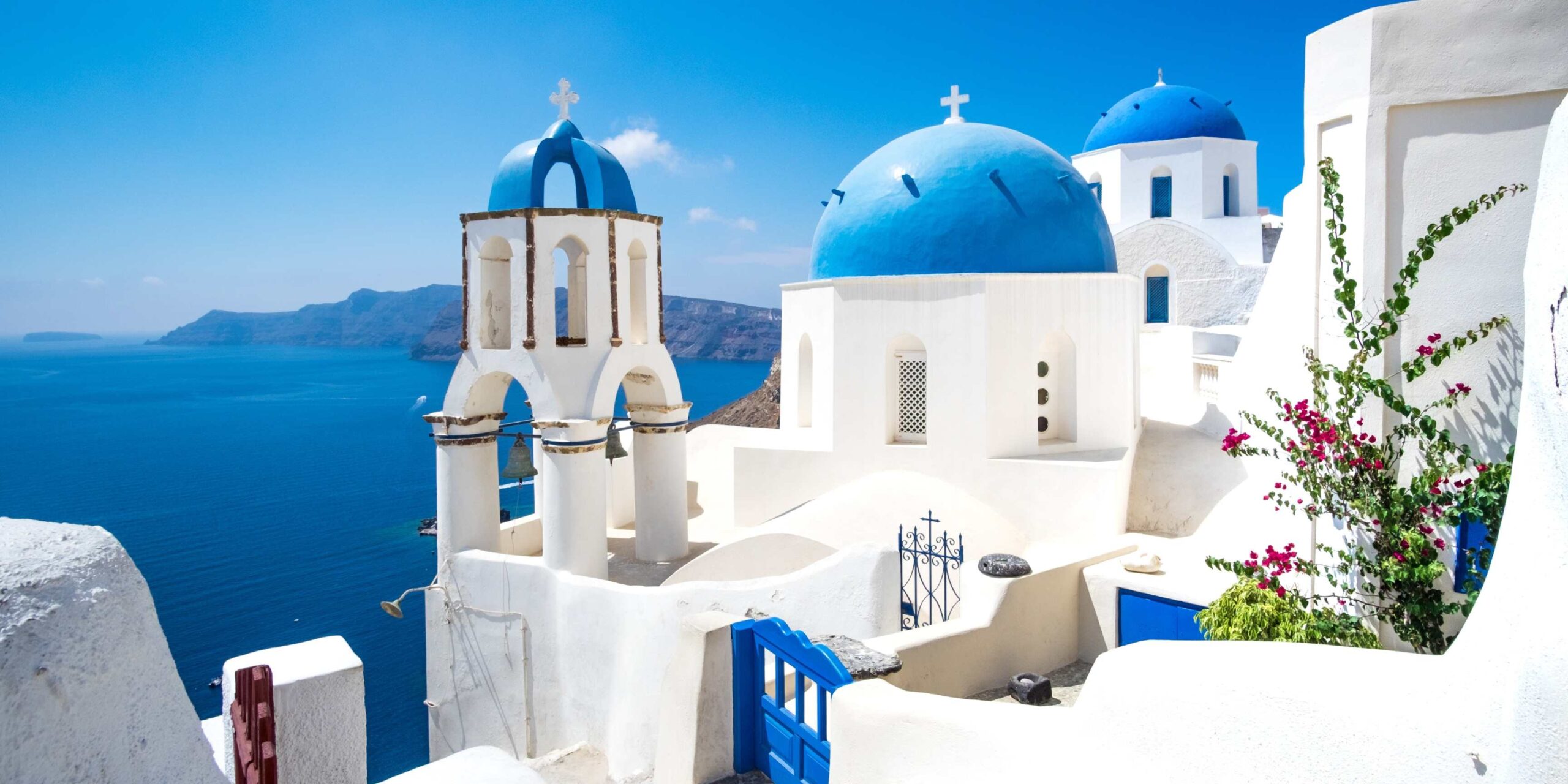 A picturesque view of Santorini featuring iconic white-washed buildings with blue domes overlooking the deep blue Aegean Sea under a clear sky.