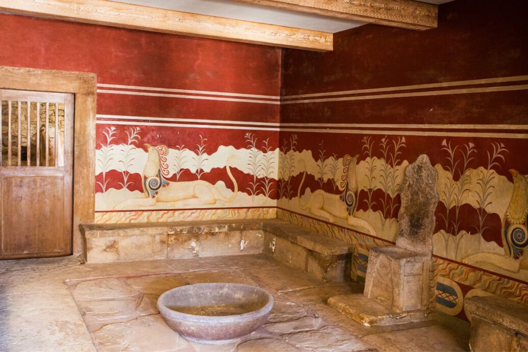 The image shows the throne room at Knossos Palace, featuring a stone throne, benches along the walls, and elaborate frescoes with red backgrounds depicting mythological creatures and foliage.