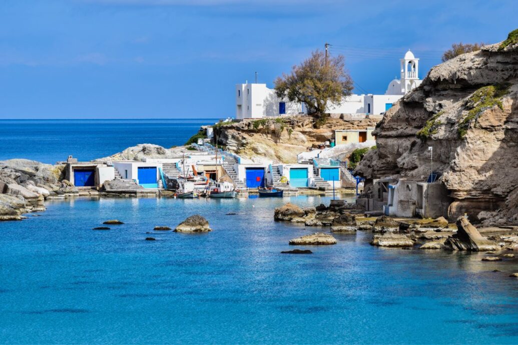 The image shows a peaceful coastal scene on Milos Island, featuring traditional white-washed buildings with bright blue doors set against rugged cliffs and calm, crystal-clear waters. Small boats are docked in the serene bay, creating a picturesque and tranquil atmosphere typical of the Greek islands.