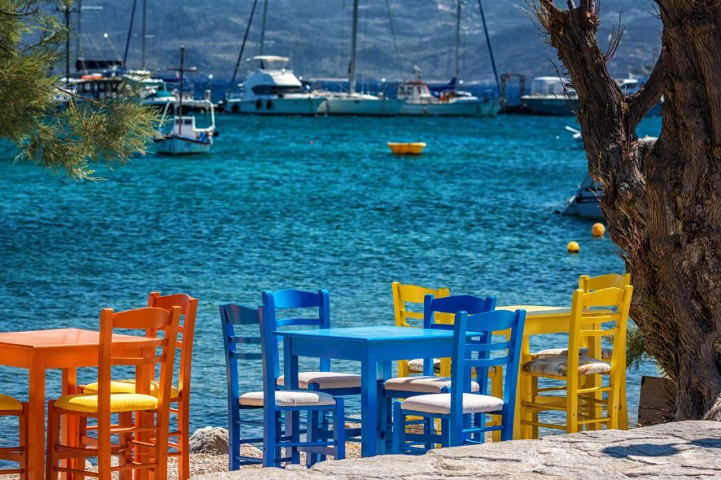 The image shows a vibrant outdoor dining area on Milos Island. The scene is filled with brightly colored tables and chairs, situated by the water's edge with boats visible in the background. The clear, turquoise water contrasts beautifully with the vibrant furniture, creating a picturesque and inviting setting typical of a Greek island.