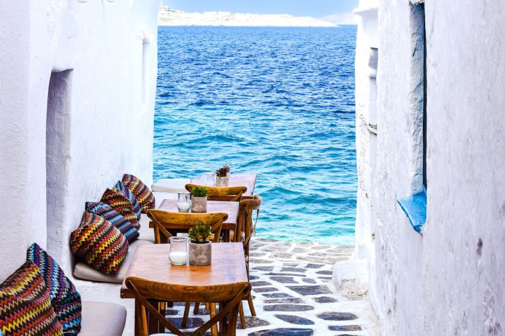 A cozy outdoor café in Mykonos, featuring colorful cushions and wooden tables, with a stunning view of the blue Aegean Sea.