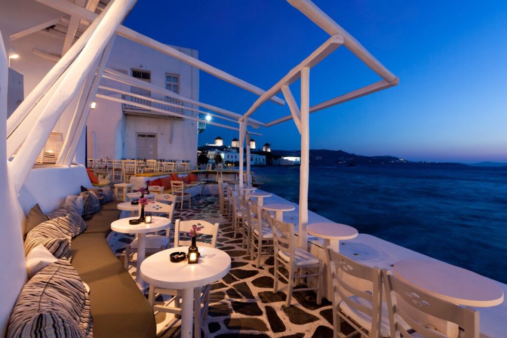 An outdoor café in Mykonos at dusk, with cozy seating and a view of the sea under a twilight sky_Mykonos Greece Vacation